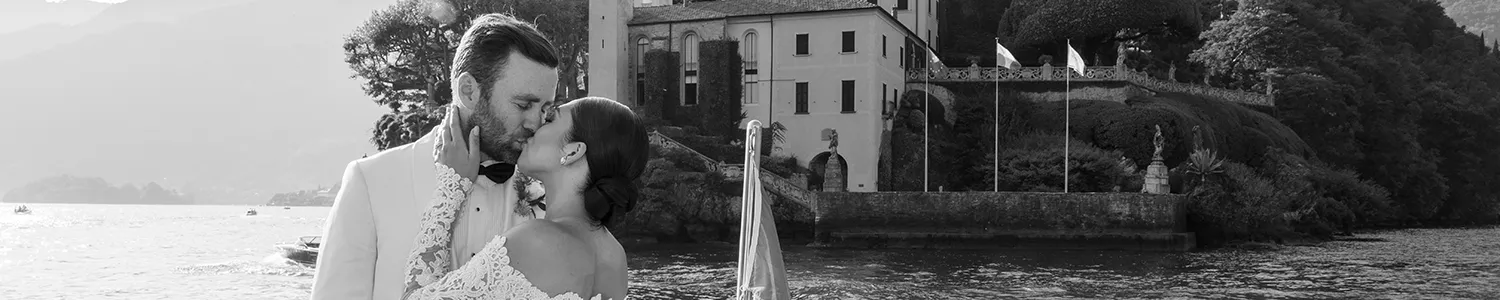 A black and white photo of a newlywed couple sharing a kiss by a scenic lake, with a classic villa and lush hillside in the background.