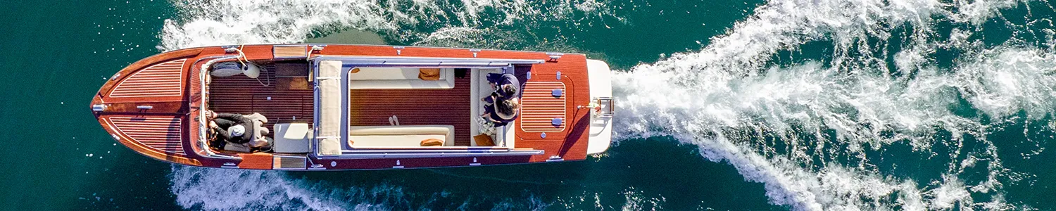 A high-end, vintage-style wooden speedboat cruising on the open sea, creating a white frothy wake in the clear blue water.