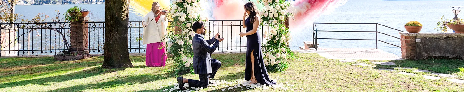 A man on one knee proposing to a woman in an elegant dress in a picturesque garden by a lake, with floral arrangements and a tree providing a romantic setting.